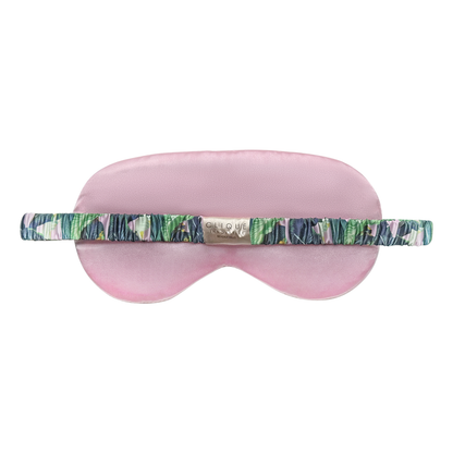 Back view of a pink satin sleep eye mask with a tropical leaf–print ruched elastic strap and a small gold “Clique Innovative Beverly Hills” brand label, shown on a white background.