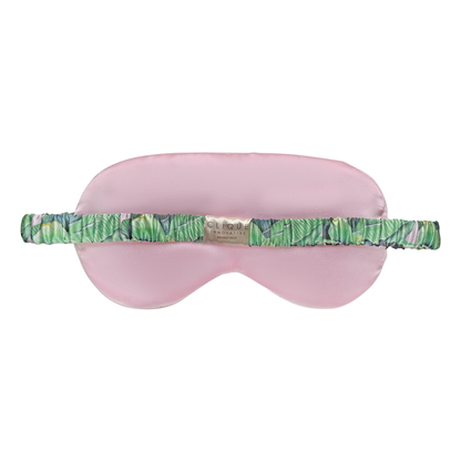 Back view of a pink satin sleep eye mask with a green tropical leaf–print elastic strap and a small “Clique Innovative Beverly Hills” brand label at the center, shown on a white background.