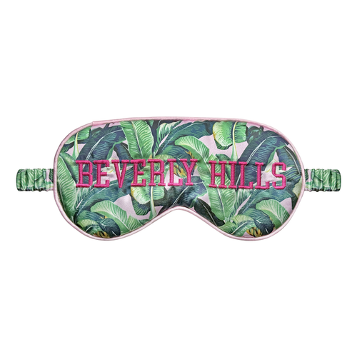 Front view of a pink-trimmed satin sleep eye mask with a green tropical banana-leaf print and BEVERLY HILLS embroidered in bright pink letters, shown on a white background.