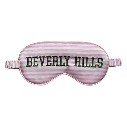 Front view of a pink-and-white striped satin sleep eye mask with “Beverly Hills” embroidered in dark green across the front, shown on a white background.