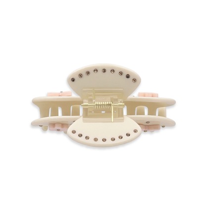 Top view of a beige claw clip showing gold rhinestones along the top edges, a gold spring mechanism, and side views of light pink flower and butterfly graphics on a white background.