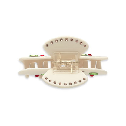 Top view of a beige claw clip showing gold rhinestones along the top edges, a gold spring mechanism, and glimpses of green leaf rhinestones and red cherry details on a white background.