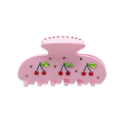 Front view of a pink claw clip with red cherry graphics with red rhinestones, green leaf accents with green rhinestones, and small gold rhinestones throughout and on the top, on a white background.