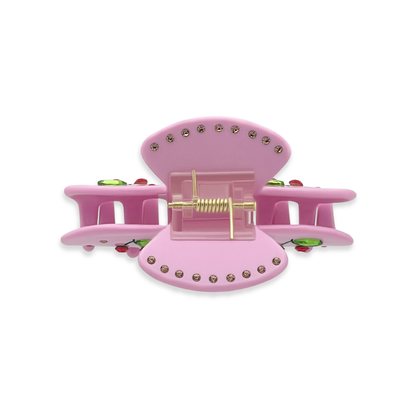 Top view of a pink claw clip showing gold rhinestones along the top edges, a gold spring mechanism, and glimpses of green leaf rhinestones and red cherry details on a white background.