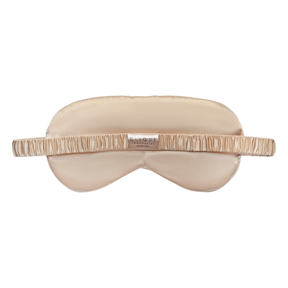 Back view of a champagne gold satin eye mask with a ruched elastic strap and a small “Clique Innovative Beverly Hills” label in the center, shown on a white background.